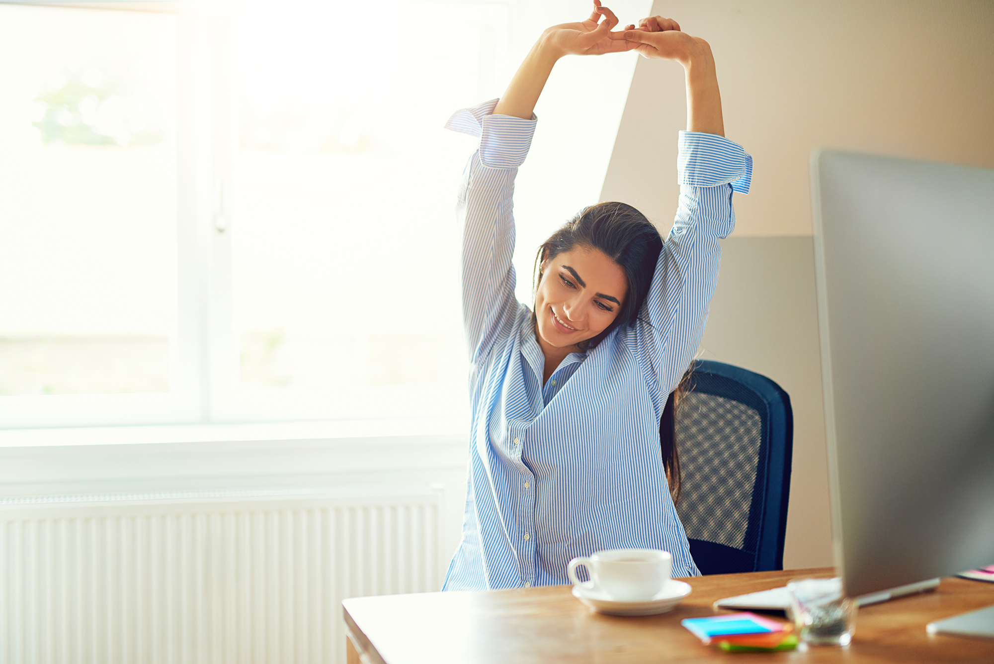 Lady in home office with computer screen, stretching in chair, cup and stationery on table, window behind her